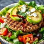 A colorful burger bowl with ground beef, fresh lettuce, diced tomatoes, pickles, shredded cheese, avocado slices, and a drizzle of burger sauce.